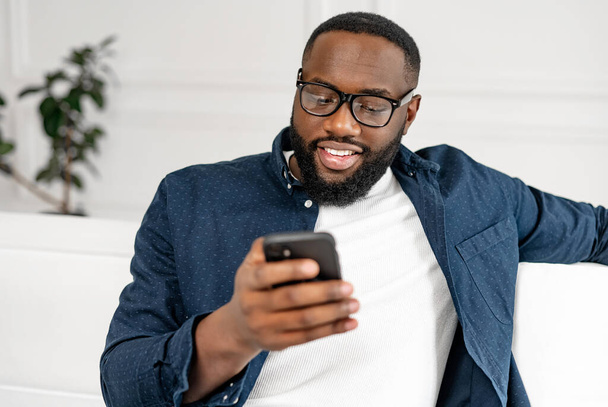 Portrait of millennial bearded African-American man in glasses using smartphone - Photo, image