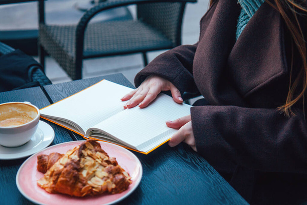 Kobieta z długimi włosami w ciemnym płaszczu, siedząca w kawiarni na ulicy z otwartym notatnikiem. Śniadanie z rogalikiem i kawą. Styl życia. Zbliżenie twarzy - Zdjęcie, obraz