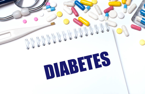 On a light background, multi-colored pills, a stethoscope, an electronic thermometer and a notebook with the text DIABETES. Medical concept. - 写真・画像