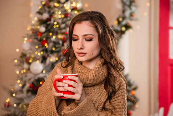 A beautiful girl in a warm sweater against the background of a Christmas tree and garlands is drinking coffee from a red mug. Model against the background of Christmas decorations - Fotó, kép