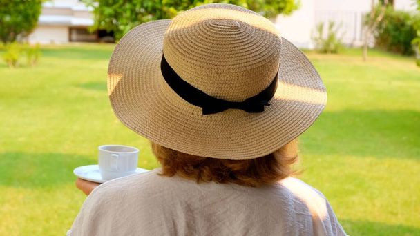 Senora femmina, 50-55 anni in un cappello di paglia tiene una tazza di bevanda calda e beve caffè, seduta al mattino su una terrazza estiva in una giornata di sole. Il concetto di buongiorno, viaggio e aspettativa di - Foto, immagini