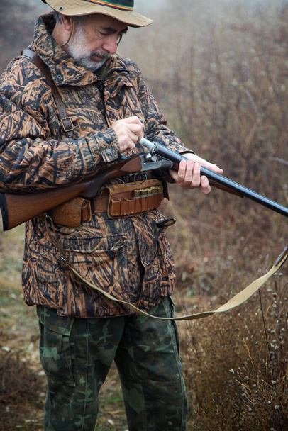 cazador hombre con barba gris en traje de cazador cartuchos de carga en un rifle de doble cañón - Foto, Imagen