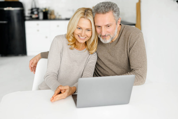 Modern cheerful mature husband and wife sit at the desk in kitchen using laptop - 写真・画像
