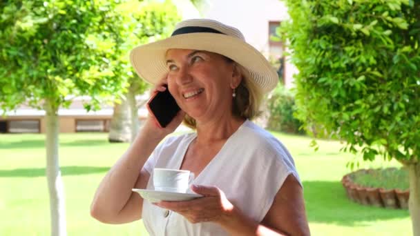 grappige blonde senora vrouw, 50-55 jaar oud, in een hoed en een witte jurk, praat met een smartphone en houdt een kopje koffie in haar handen op haar terras op een zonnige ochtend op een zomerse dag. Freelance - Video