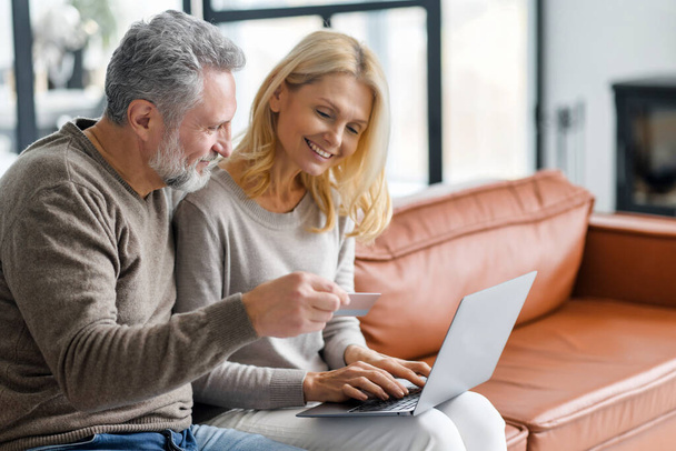 Cheerful middle-aged couple using laptop for making purchases in online shopping - Foto, imagen