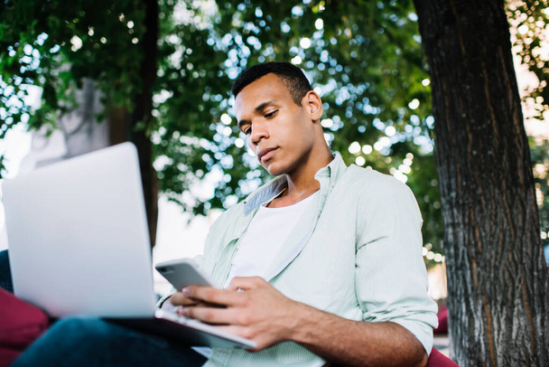 Low angle of confident young black man in casual outfit sitting on bag chair and working remotely on netbook while messaging on smartphone in street near trees in summer day - 写真・画像