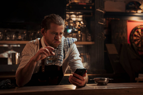 白人男性バーテンダー一人で座っていますバーでテキストメッセージ上の携帯電話デバイスながらウイスキーを飲み - 写真・画像