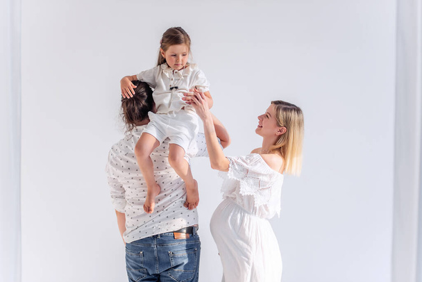 Young modern family have fun together in white loft interior. Mother is pregnant, father holds daughter in arms, hugs his wife. A man with glasses and a beard. Blond barefoot woman. Healthy pregnancy - Foto, Imagen