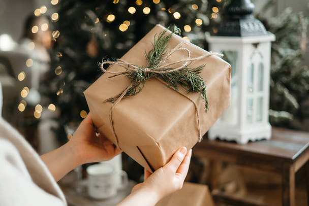 Fókuszálj a csomagolt karácsonyi ajándékra, gratulálj a téli ünnepekhez, emlékeztetve az ajándékok elkészítésére, pózolj a díszített fa közelében. - Fotó, kép
