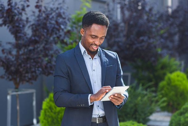 Jeune homme d'affaires afro-américain en costume d'affaires formelle debout travaillant avec une tablette dans les mains sur fond immeuble de bureaux moderne à l'extérieur. Homme utilisant smartphone ou téléphone portable en plein air rue de la ville - Photo, image