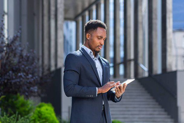 Young african american businessman in formal business suit standing working with tablet in hands on background modern office building outside. Man using smartphone or mobile phone outdoors city street - Фото, зображення