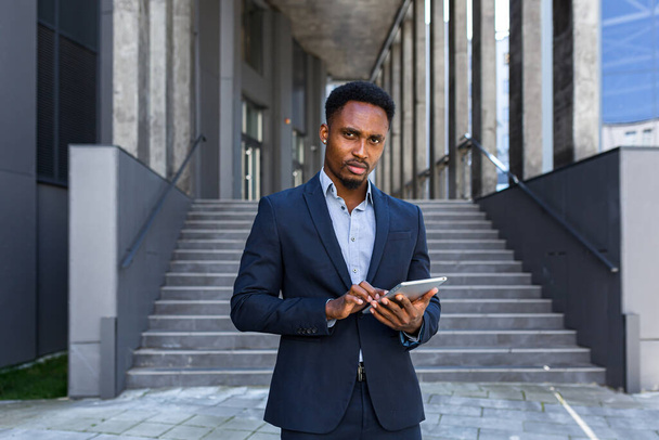 Young african american businessman in formal business suit standing working with tablet in hands on background modern office building outside. Man using smartphone or mobile phone outdoors city street - Valokuva, kuva