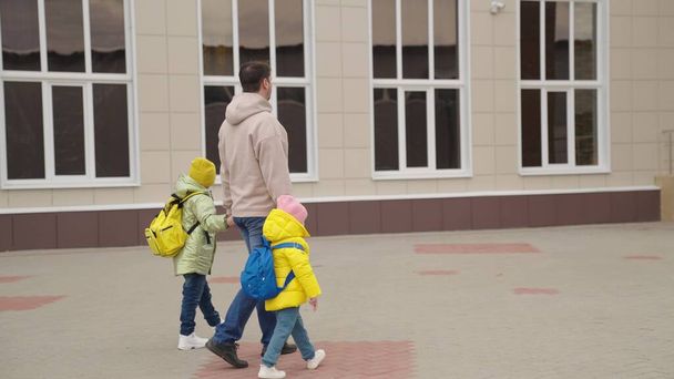Father leads little children with backpacks holding hand to school, childhood schoolyard, dad and daughters rush to class together, happy family, dad took kid after school day, preschool education - Foto, immagini