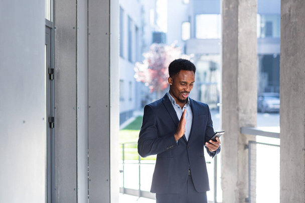 African American businessman grinds on video call uses phone and app, outside office, cheerful and successful man in business suit - Fotoğraf, Görsel