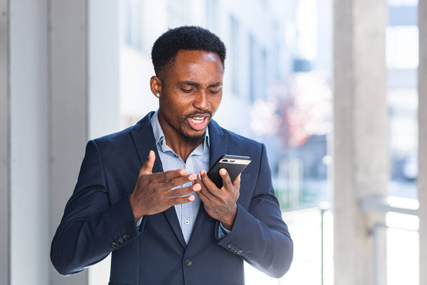 African American male businessman angry talking on mobile phone outside. Business man having phone call outdoors. Nervous mad professional conversation on smartphone. Irritated speak at urban street - Foto, Imagem