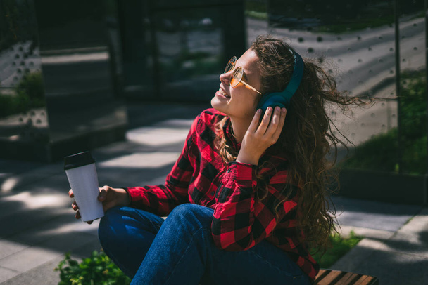 Cheerful smiling girl listens to music on headphones with coffee in the city park on a bench. - Foto, immagini