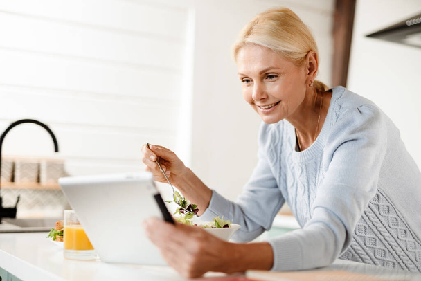 Усміхнена жінка середнього віку, що тримає планшетний комп'ютер, спираючись на кухонну лавку зверху
 - Фото, зображення