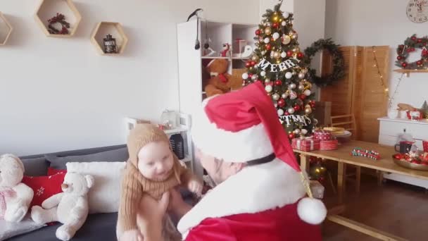 Санта-Клаус в красно-белом наряде играет с малышом на фоне украшенных елок с сверкающими гирляндами и коробками подарков - Кадры, видео