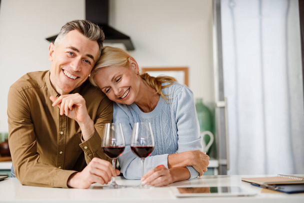 Smiling middle aged couple standing together at the kitchen drinking red wine at home - 写真・画像