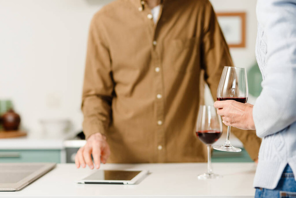 Middle aged couple standing together at the kitchen drinking red wine at home using tablet, cropped - Photo, Image