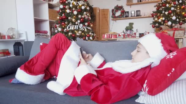 Fiatal Mikulás piros-fehér öltönyben fekszik a kanapén a szilveszteri dekoráció hátterében, és gratulál a telefonhoz. - Felvétel, videó