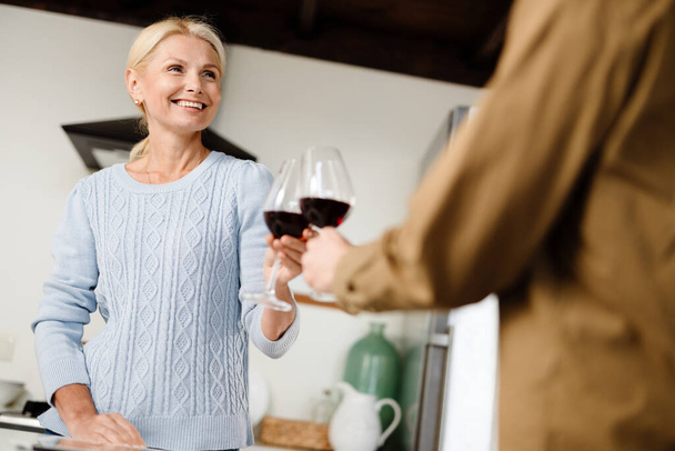 Smiling mid aged couple drinking red wine in kitchen at home - Φωτογραφία, εικόνα