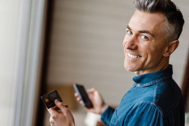 Smiling mid aged white man holding mobile phone and credit card standing in the room - Fotoğraf, Görsel