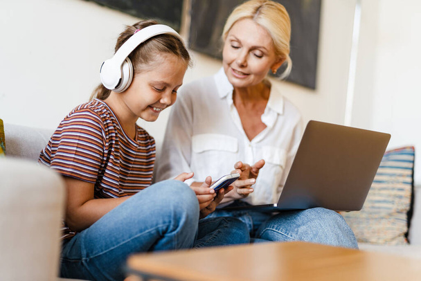 White mother and daughter using cellphone and headphones while sitting on couch at home - Foto, Imagen