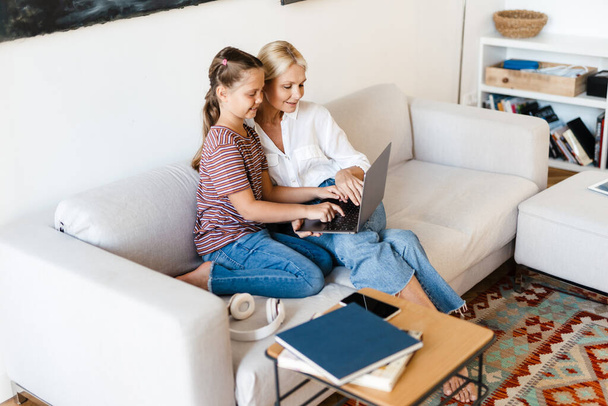 Bílá matka a dcera s úsměvem a pomocí mobilu, zatímco sedí na gauči doma - Fotografie, Obrázek