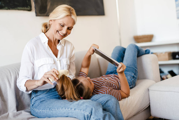White girl using tablet computer while her mother combing her hair at home - Zdjęcie, obraz