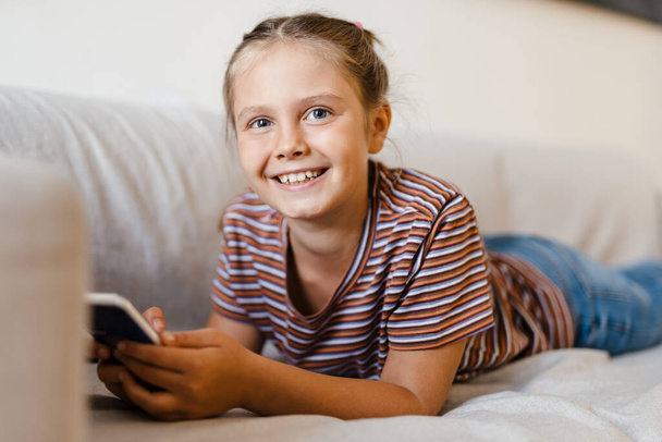 White preteen girl smiling and using mobile phone while lying on couch at home - Photo, Image