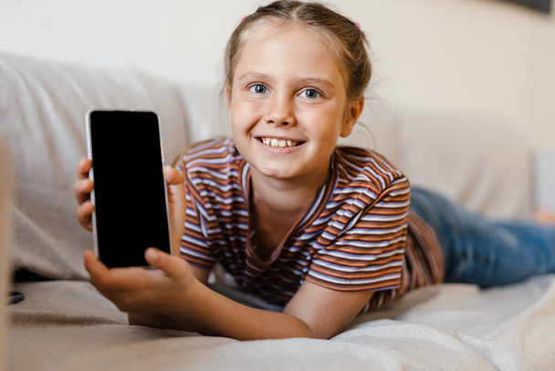 Bianco preteen ragazza sorridente e mostrando il cellulare mentre sdraiato sul divano a casa - Foto, immagini