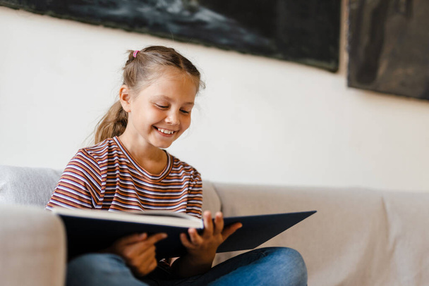 White preteen girl smiling and reading book while sitting on couch at home - Фото, зображення
