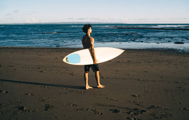 Side view full body faceless fit shirtless male surfer carrying white surfboard and walking on sandy beach towards waving sea during clear day - 写真・画像