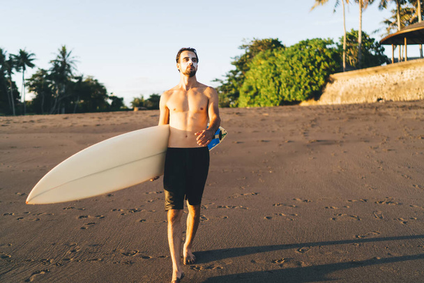 Confident young shirtless male traveler walking along empty sandy beach with surfboard and looking away while enjoying sunny day in tropical resort - 写真・画像