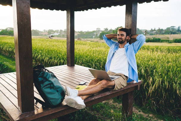 Young content bearded self employed man with netbook and hands behind head sitting with crossed legs near backpack in wooden alcove and looking away - Foto, Imagem