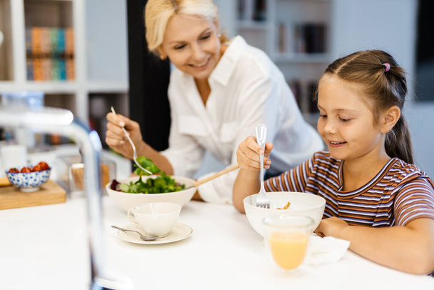 White mother and daughter smiling while having lunch in kitchen at home - Φωτογραφία, εικόνα