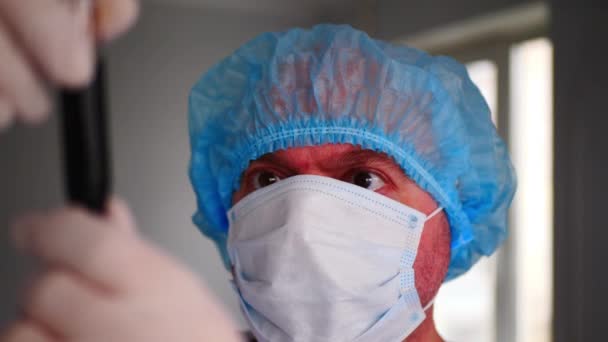 Профессиональный врач осматривает пробирку крови в лаборатории. Медсестра в медицинской хирургической одноразовой маске для лица и синей медицинской шапке осматривает стеклянную пробирку с красной жидкостью в больнице - Кадры, видео