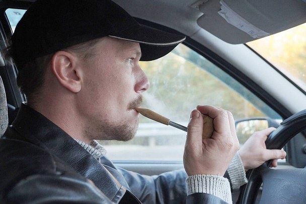 een jonge man rookt een pijp tijdens het besturen van een auto, zijaanzicht - Foto, afbeelding