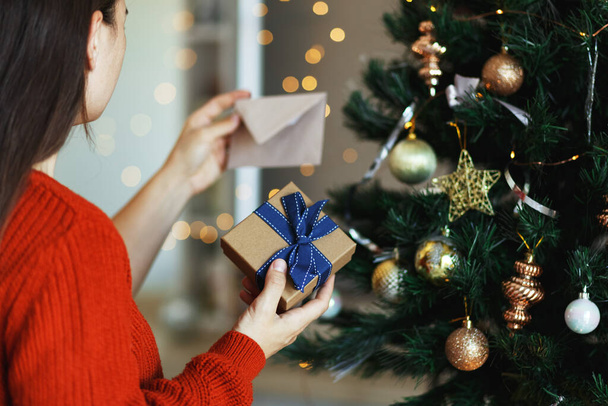 Mujer abriendo caja de regalo junto al árbol de Navidad y leyendo la tarjeta de felicitación. Navidad, vacaciones de Año Nuevo  - Foto, imagen