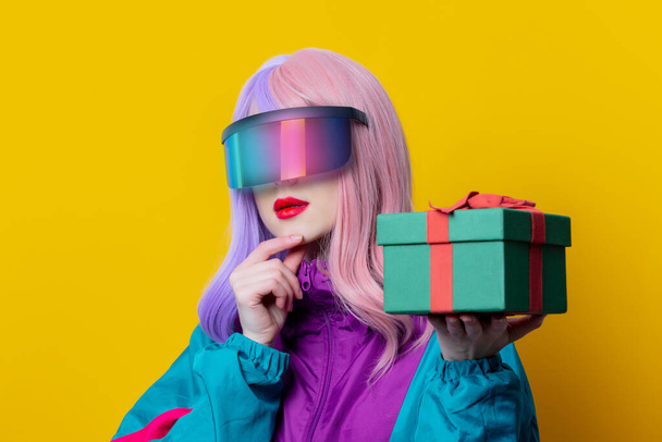 VR gözlüklü ve 90 'ların spor takımlı stil kadını sarı arka planda hediye kutusu tutuyor. - Fotoğraf, Görsel