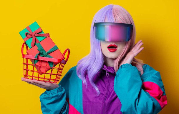 Style woman in VR glasses and 90s sport suit hold basket with gift boxes on yellow background - Photo, Image