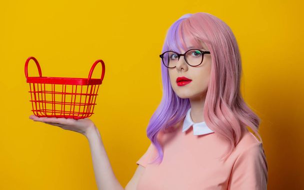 Chica con pelo morado y vestido rosa con cesta de la compra roja sobre fondo amarillo - Foto, Imagen