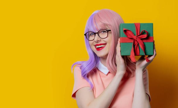 Girl with purple hair and pink dress hold gift box on yellow background - Фото, изображение