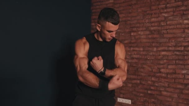 Jovem homem musculoso em uma camiseta sem mangas preto fica contra um fundo de parede de tijolo vermelho e demonstra bíceps grande - Filmagem, Vídeo