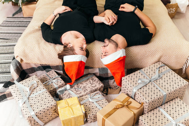 Υπέροχο νεαρό ρομαντικό ζευγάρι ξαπλωμένο κρεβάτι κοντά δώρα παρουσιάζει γιρλάντες σπίτι ζεστή εσωτερική ατμόσφαιρα Νέο Έτος καπέλο Χριστουγεννιάτικο δέντρο διακοσμήσεις διακοπές κόμμα γιορτάζει έννοια χειμωνιάτικο βράδυ  - Φωτογραφία, εικόνα
