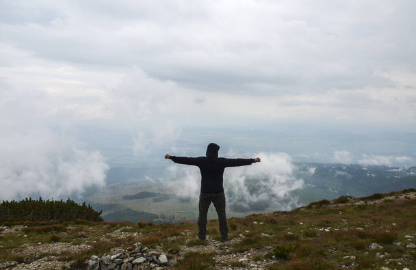 De wandelaar staat met opgeheven handen op een bergtop en geniet van het uitzicht op de bewolkte vallei. Achteraanzicht. Buitenactiviteiten, lifestyle concept - Foto, afbeelding