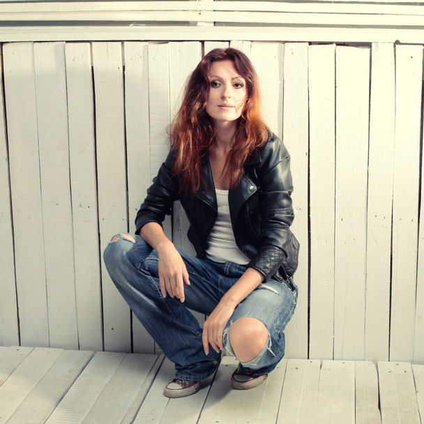 Beautiful young woman in jeans sitting against grunge wood wall - Photo, image