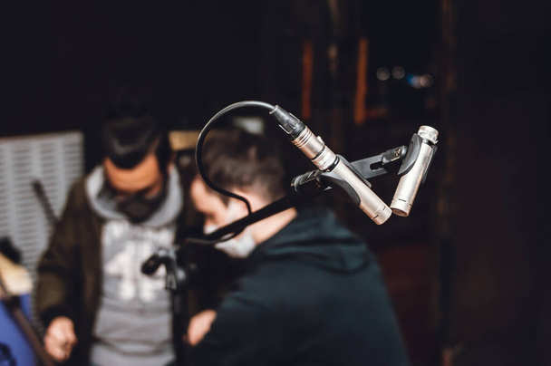 dwa mikrofony kondensacyjne zamontowane na statywie, w żywopłocie nagraniowym studia muzycznego, z dwoma rozmazanymi osobami w tle, koncepcja przemysłu muzycznego. - Zdjęcie, obraz