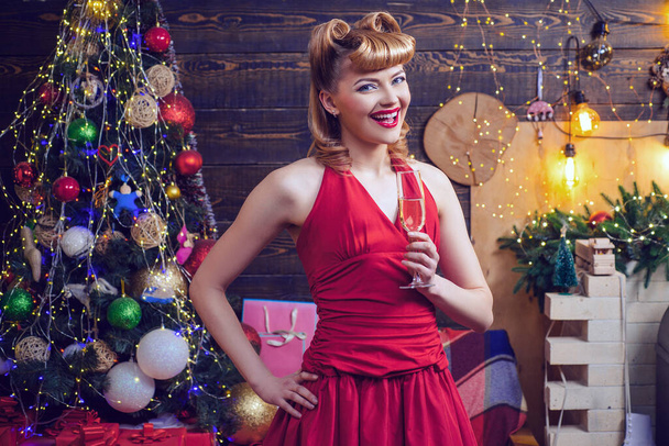 Smiling ponup woman in evening red dress with glass of sparkling wine over home Christmas tree background. Fashion and beauty make-up and hairstyle for Christmas. - 写真・画像